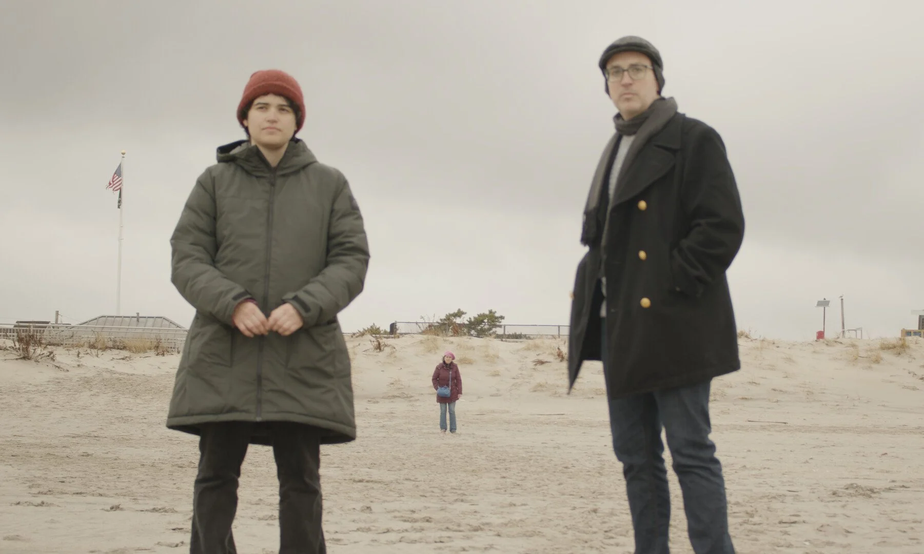 Two men standing on a sandy beach with gray clouds overhead and a woman standing in the background. One man wears a red beanie and green jacket, the other wears glasses, a black coat, and a scarf.