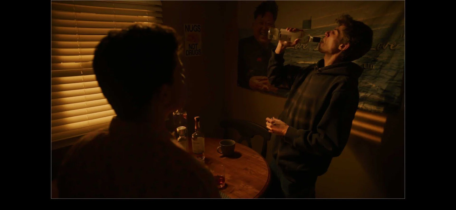 Three people in a dimly lit room, one sitting at a table and two standing behind; one standing man is drinking from a bottle, and a poster on the wall with the text 'Nugs Not Drugs' is visible.