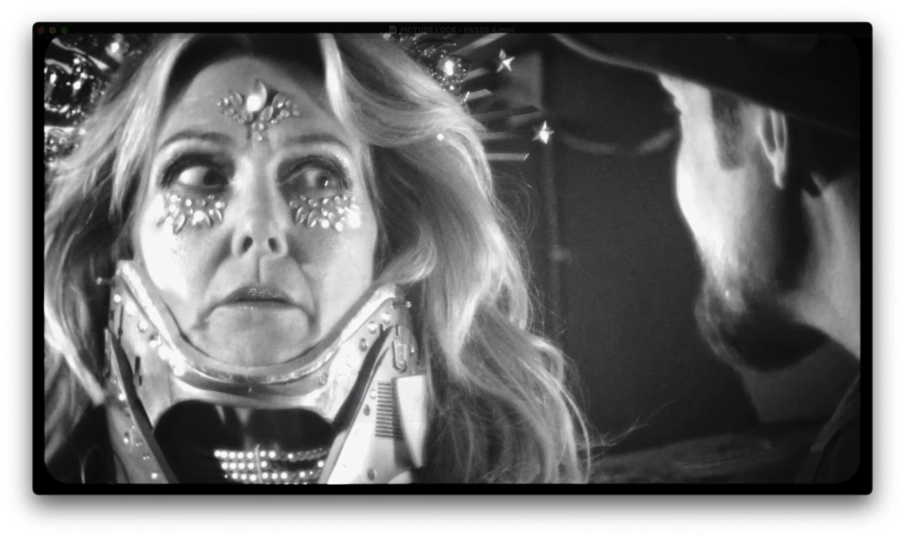 A woman with elaborate face makeup and pasties looking scared, facing a person wearing glasses and a hat in a dimly lit setting.