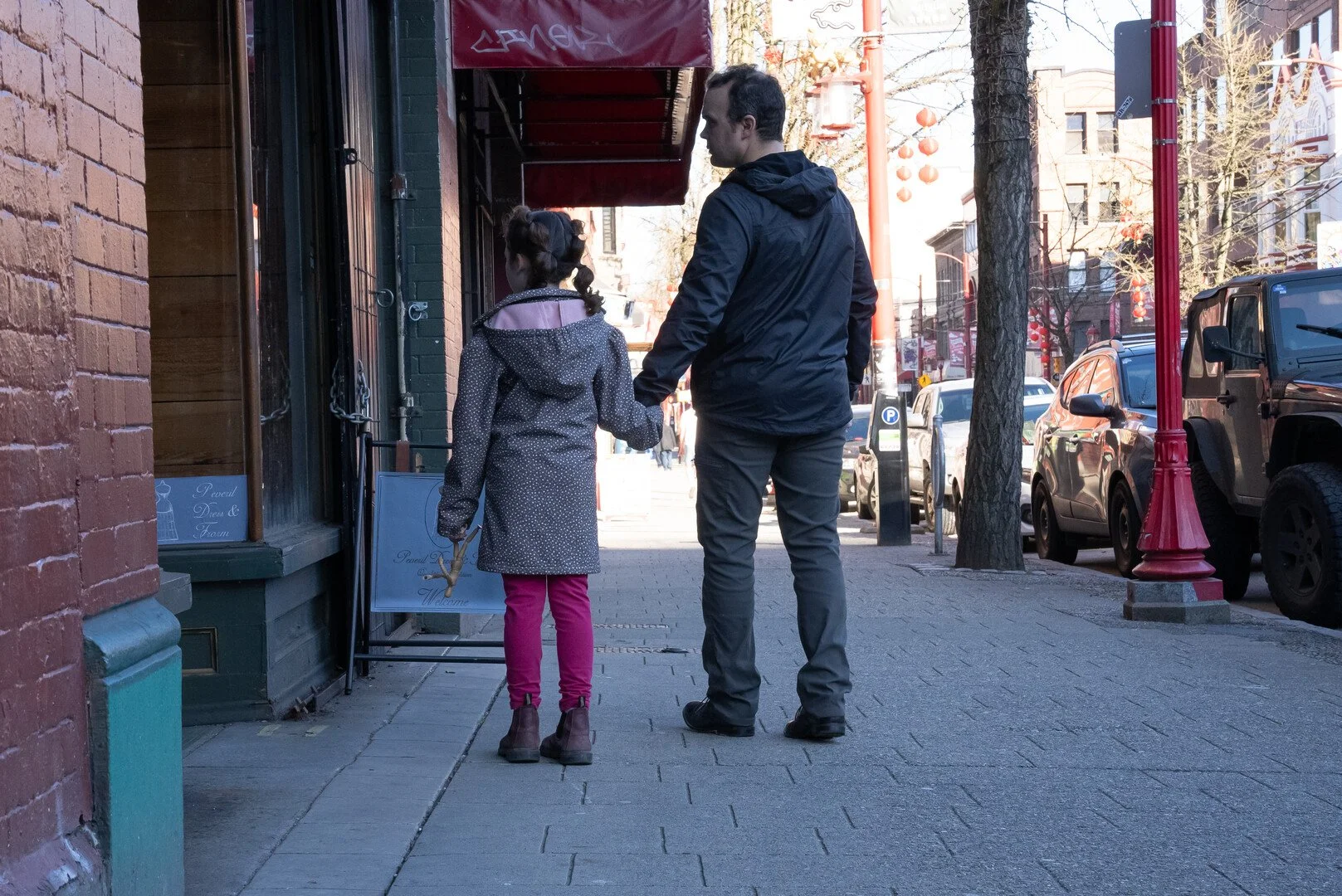 A man and a young girl holding hands walk down a city sidewalk past a storefront with a red awning, parked cars, and trees lining the street.