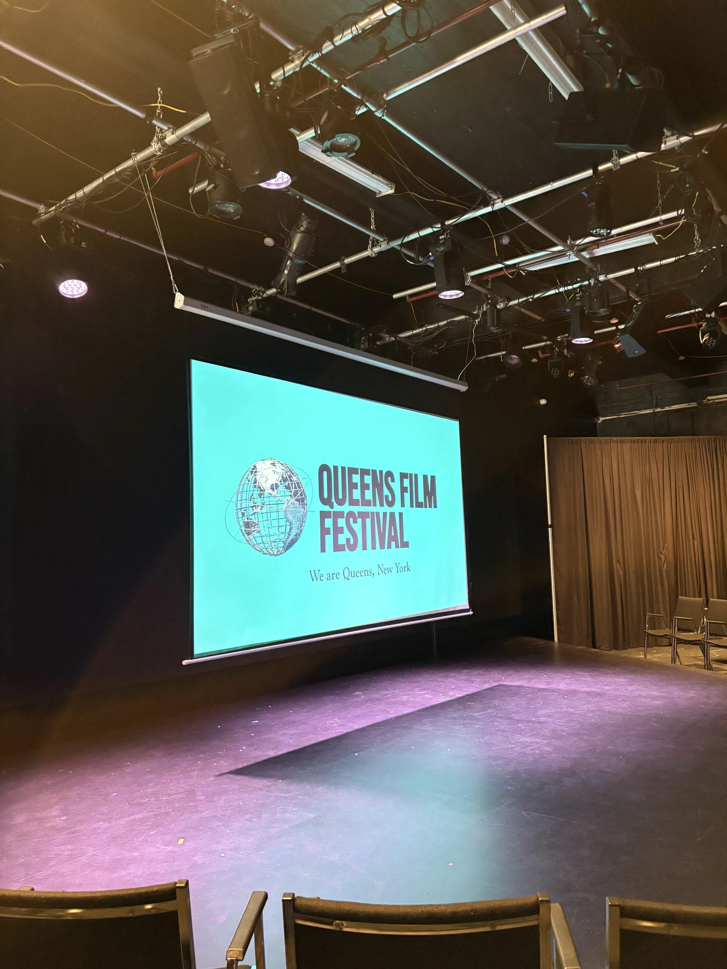 Stage with a large screen displaying Queen's Film Festival logo and text, chairs arranged on the side, spotlights and lighting equipment overhead in a black-box theater setting.