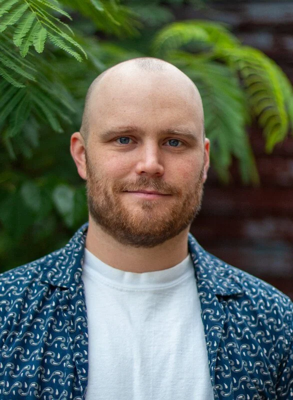 A man with a bald head and beard, wearing a white t-shirt and a blue patterned button-up shirt, standing outdoors with green plants and trees in the background.