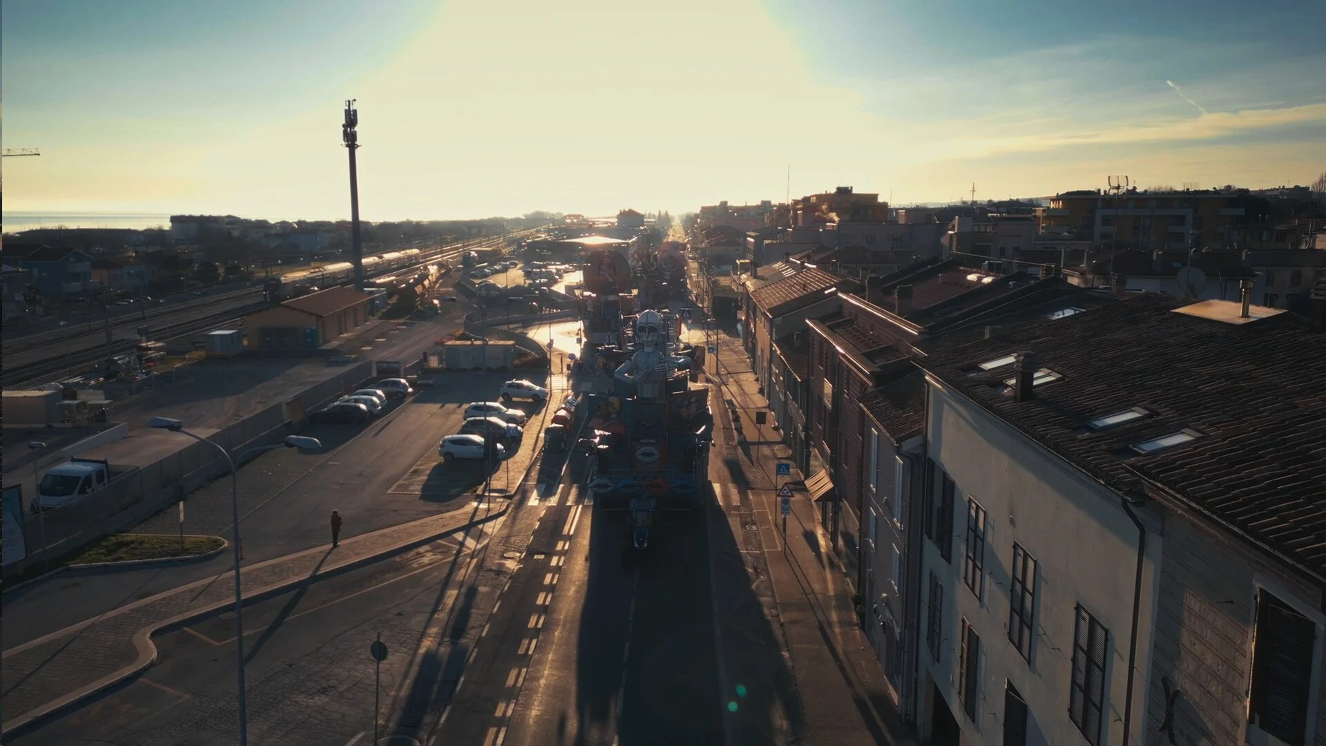 An aerial view of a city street during sunset showing parked cars, buildings, and a large figure made of flowers or artificial materials in the center of the street.
