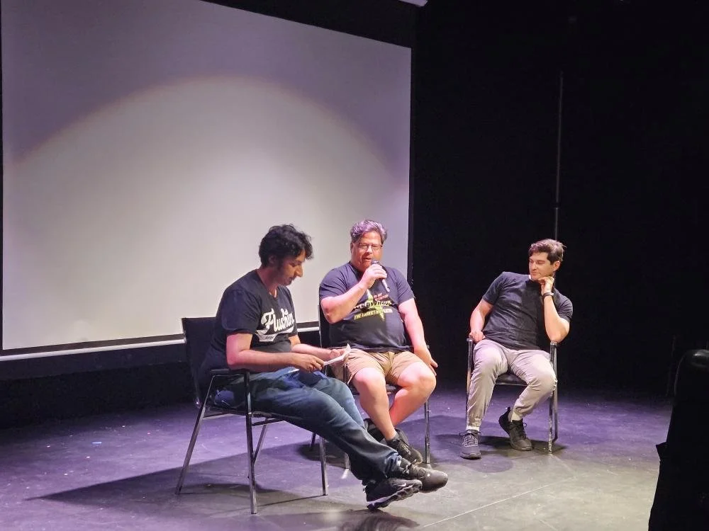 Three men sitting on chairs on a stage, with a blank projection screen behind them. One man is speaking into a microphone, while the other two are listening.