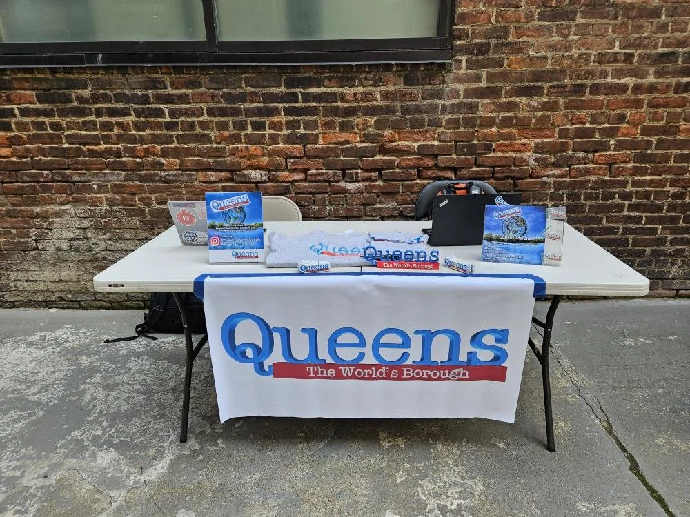 Table with Queens-themed merchandise and promotional materials, set up outdoors against a brick wall, with a large Queens banner hanging on the front of the table.