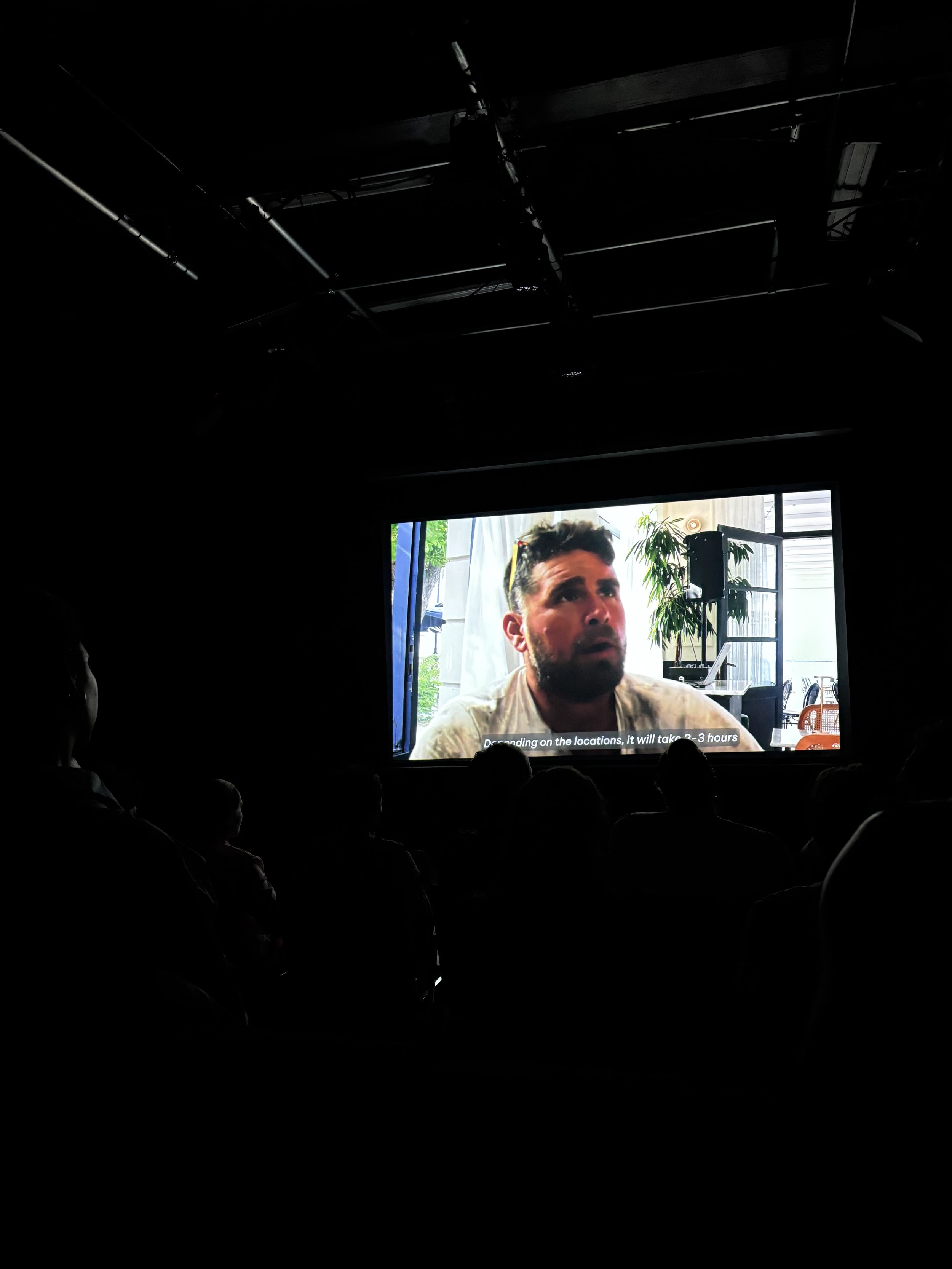 Audience watching a large screen displaying a man speaking in an indoor setting with plants and windows in the background.