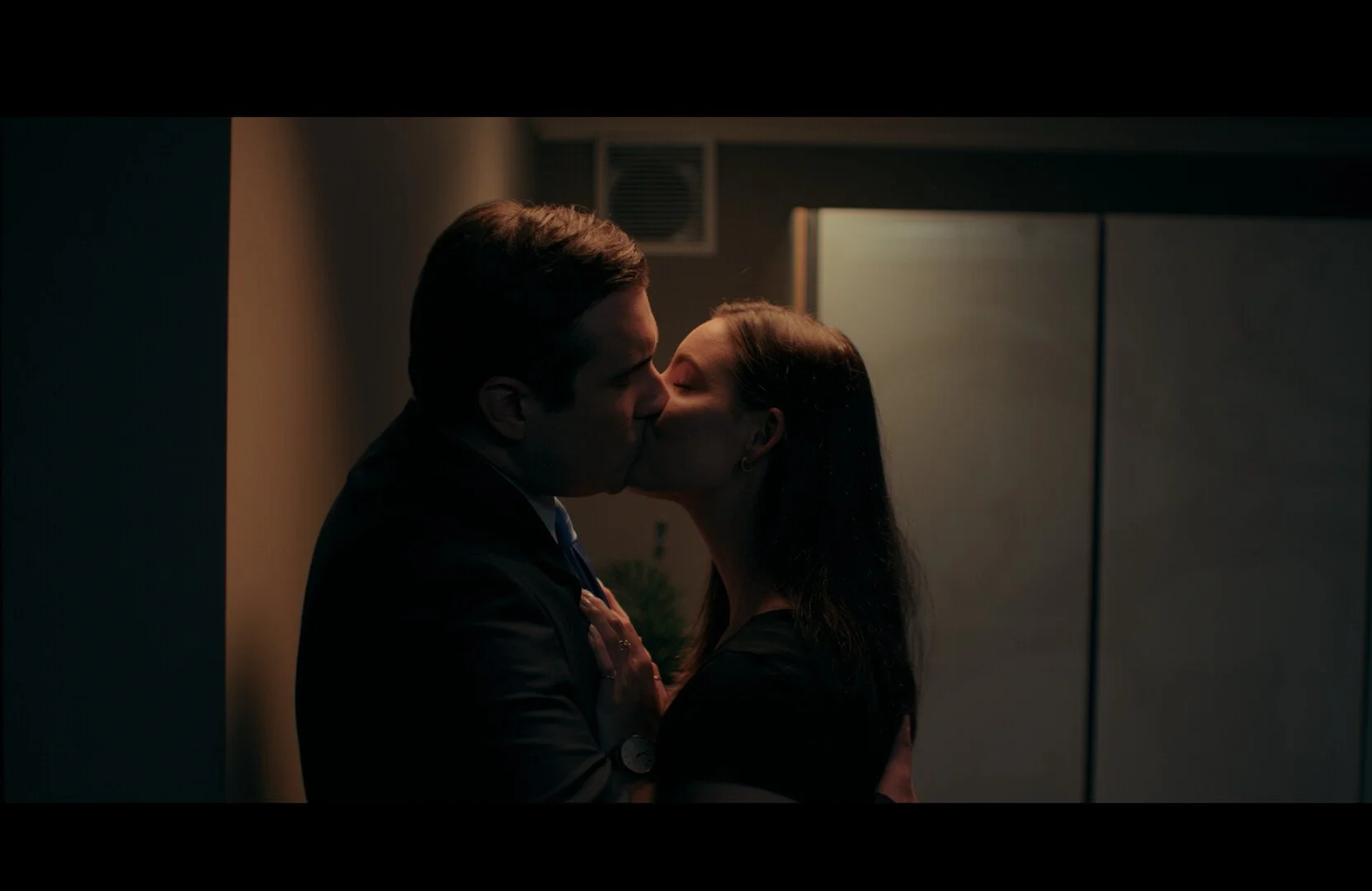 A man and woman kissing in a dimly lit room, with the man dressed in a suit and the woman with dark hair, behind a blue partition.