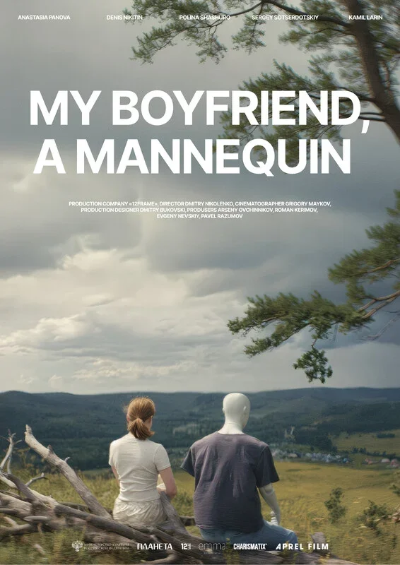 A woman with light brown hair wearing a white shirt sitting on a log, and a mannequin dressed in a dark shirt sitting on a log in a grassy field with a cloudy sky and trees in the background.