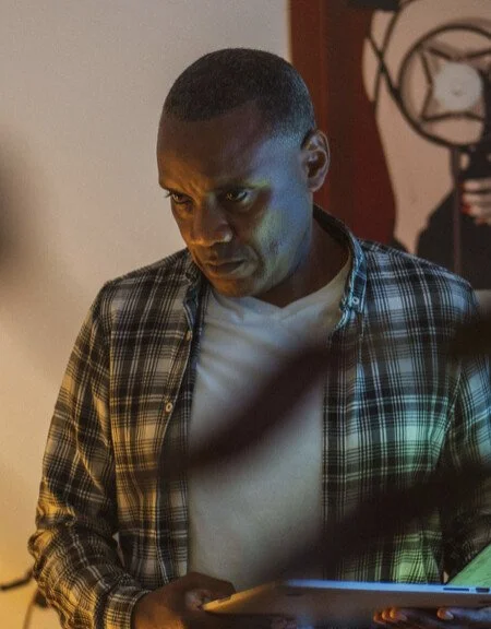 A man with short hair wearing a plaid shirt over a white t-shirt, looking down at a tablet or laptop.