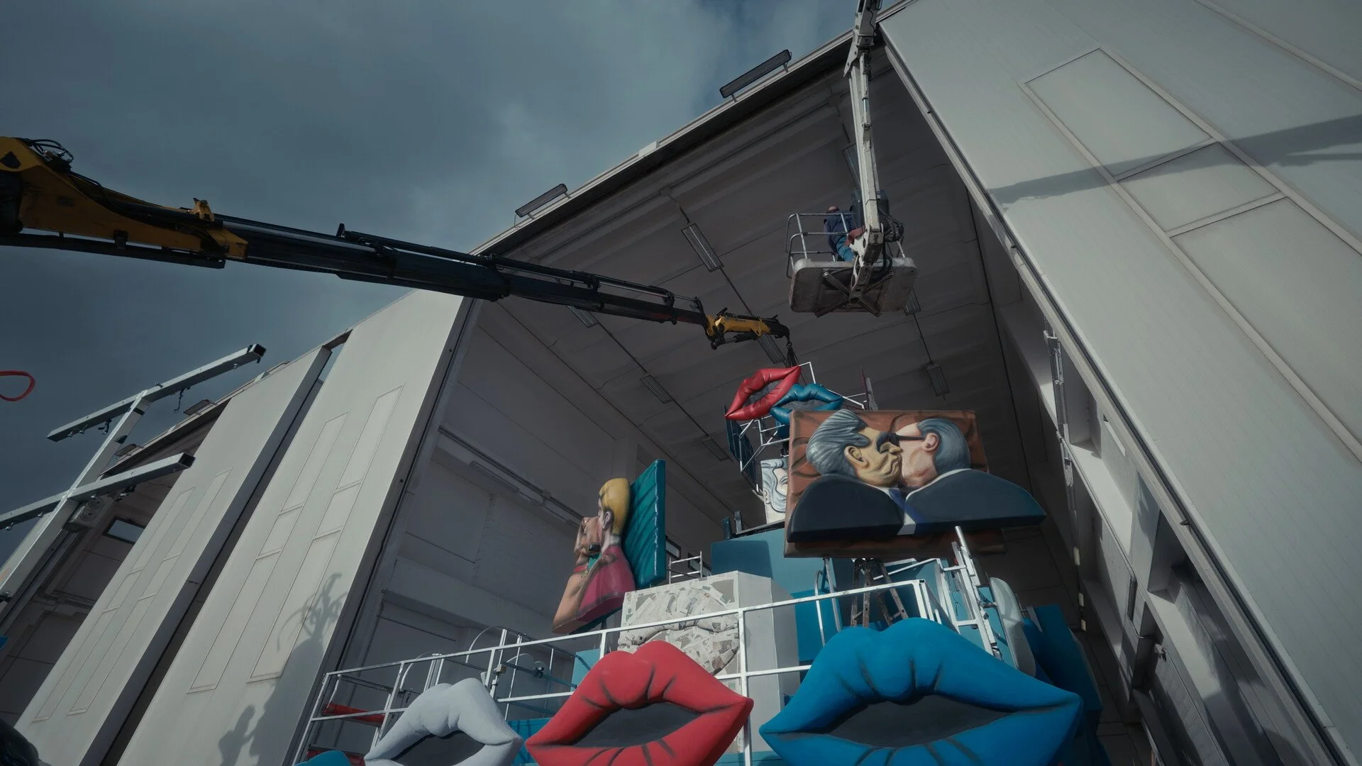 A crane lifting large, colorful sculptures and paintings, including lips, faces, and abstract art, inside an industrial warehouse with a cloudy sky visible above.
