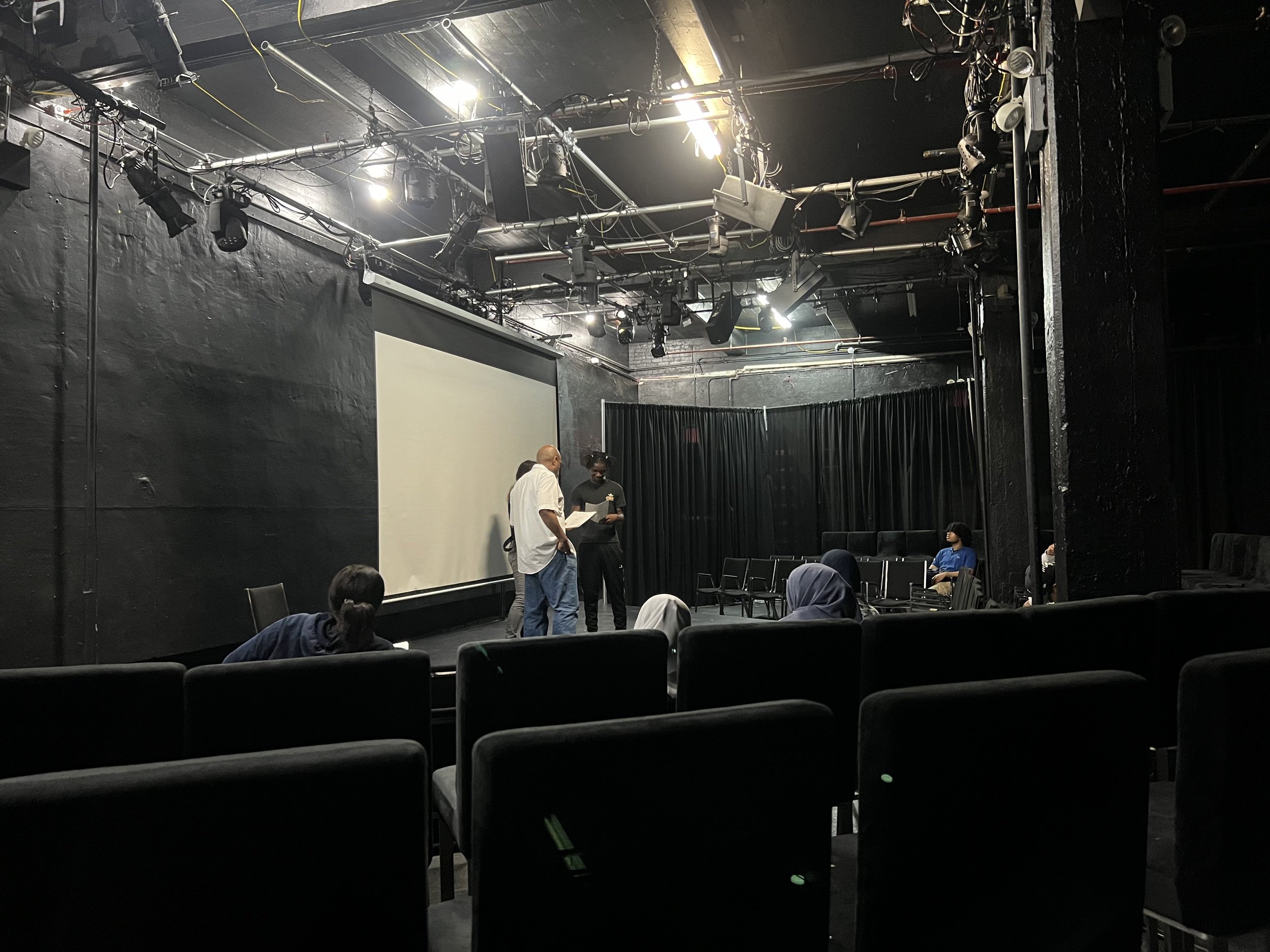 An empty black-box theater or performance space with a few people on stage, some seated in the audience area, and a large projection screen on the wall.