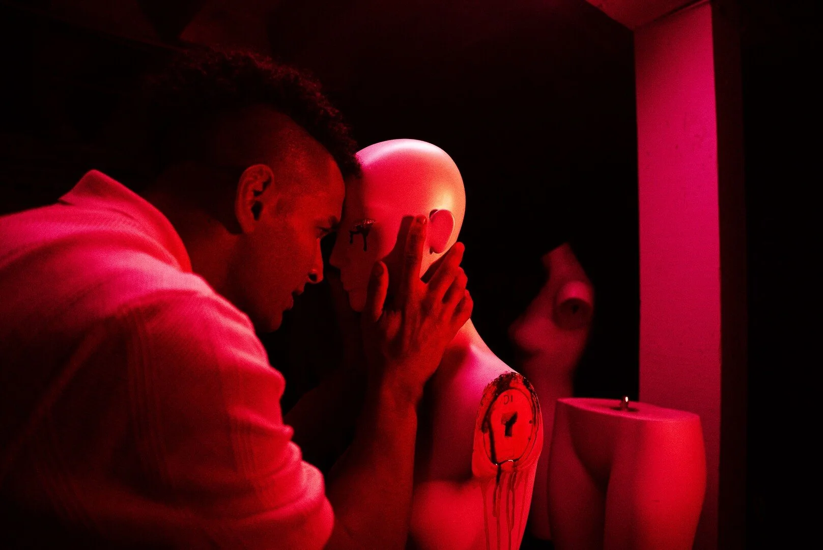 A person with a mohawk hairstyle leaning over a mannequin holding a clock to its face in a dark room illuminated by red light.