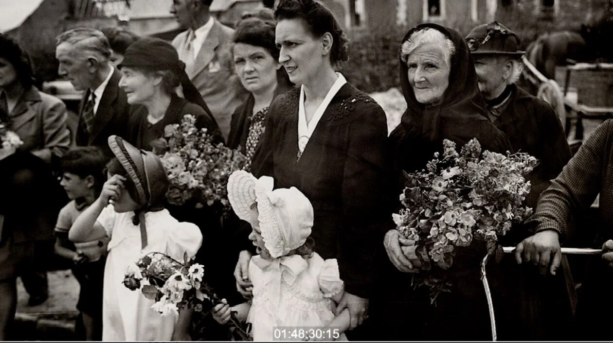 Black and white photo of a group of people at a gathering or procession, with women holding flowers and children wearing hats, outdoors in a residential area.