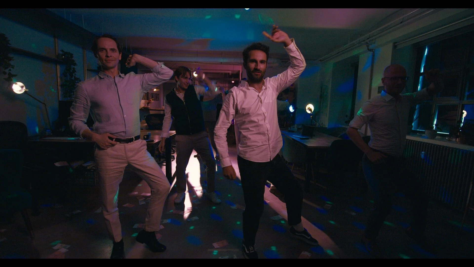 People dancing at a party in a dimly lit room with colorful lights and windows