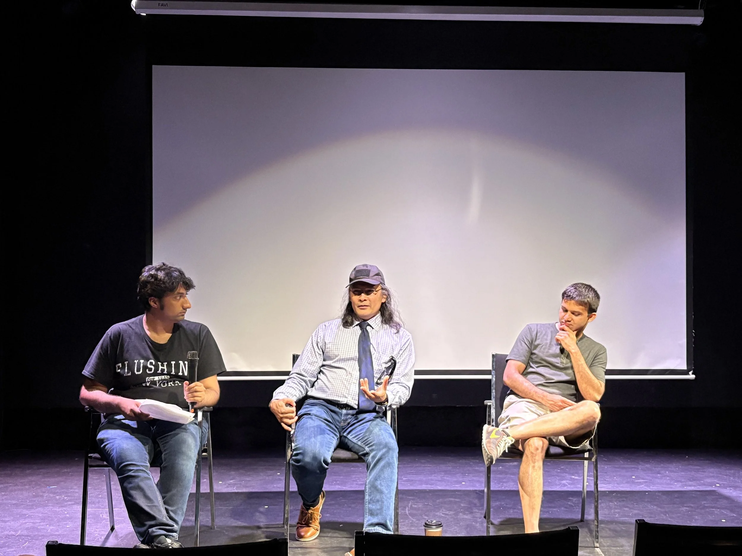 Three actors sitting on chairs on stage, engaged in a discussion or performance. The person in the middle is speaking, wearing glasses, a hat, and a tie. The stage has a large blank screen behind them.