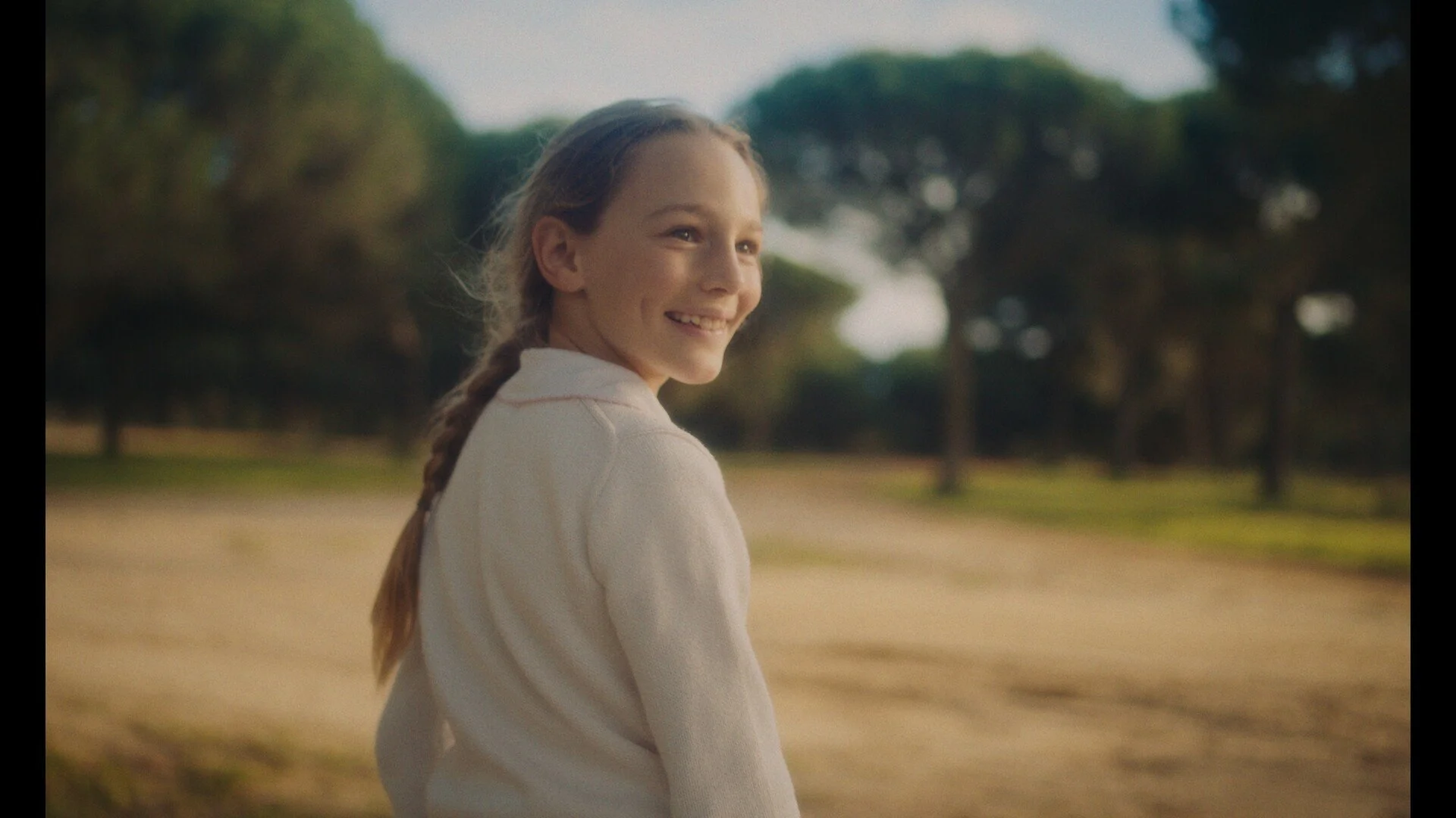 A young girl with a braid smiling and looking over her shoulder outdoors in a park with trees and a dirt path.