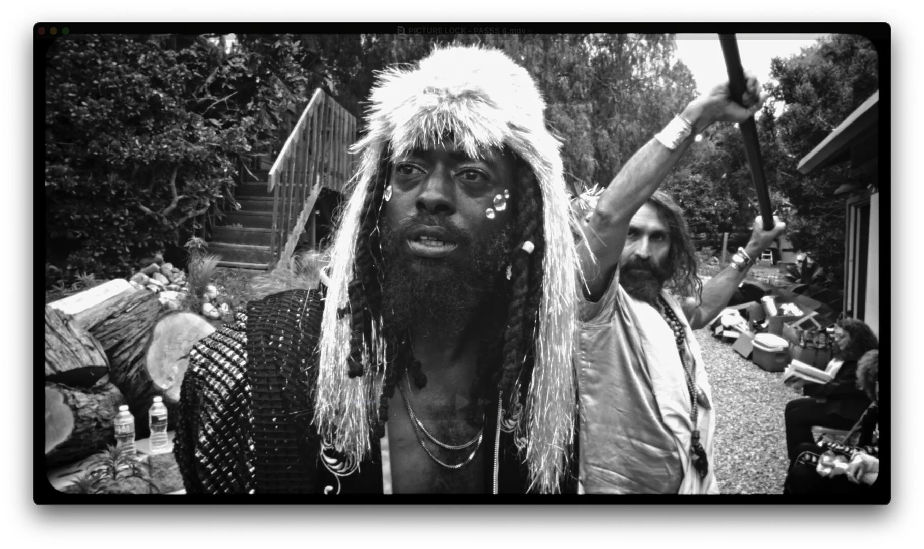 Two men outdoors, one wearing a large fur hat and braided hair, the other with long hair and a beard, holding a stick. People are sitting on benches in the background.