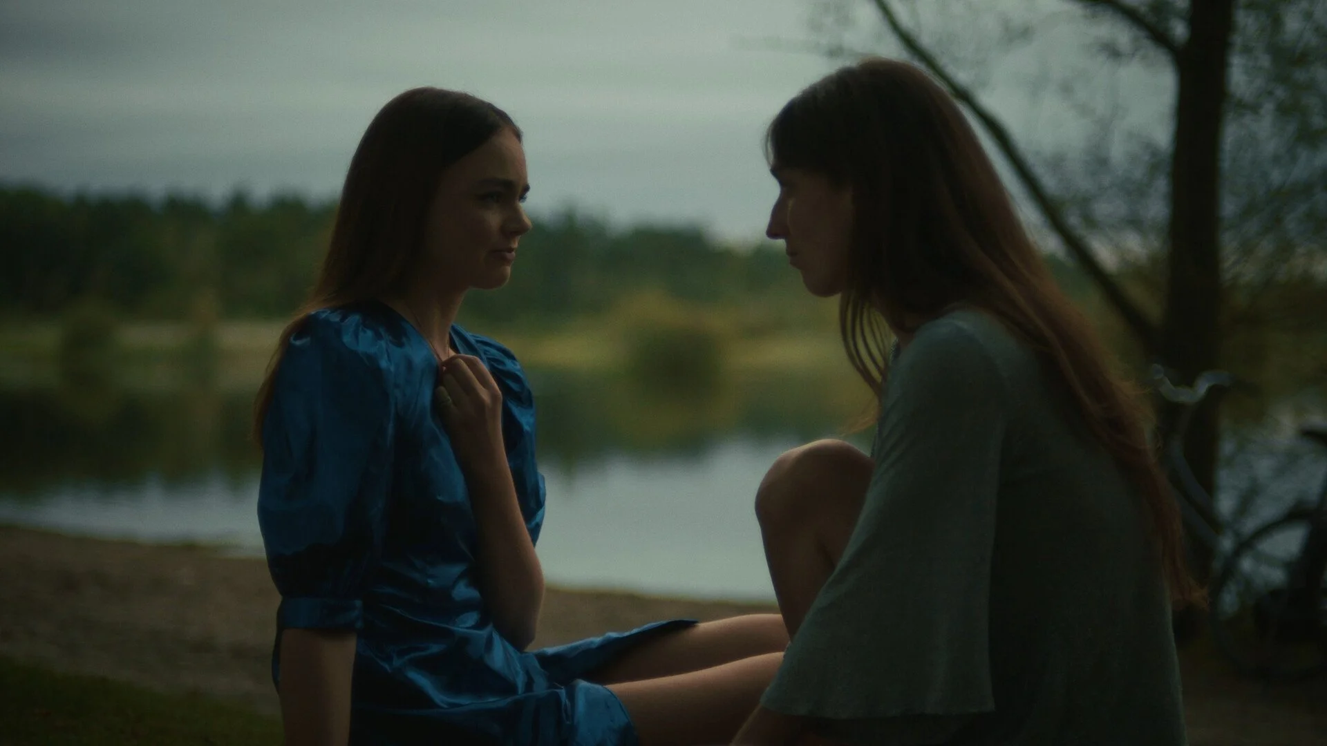 Two women sitting near a river in a natural setting, facing each other, with one woman holding the other’s hand. It appears to be dusk or dawn.