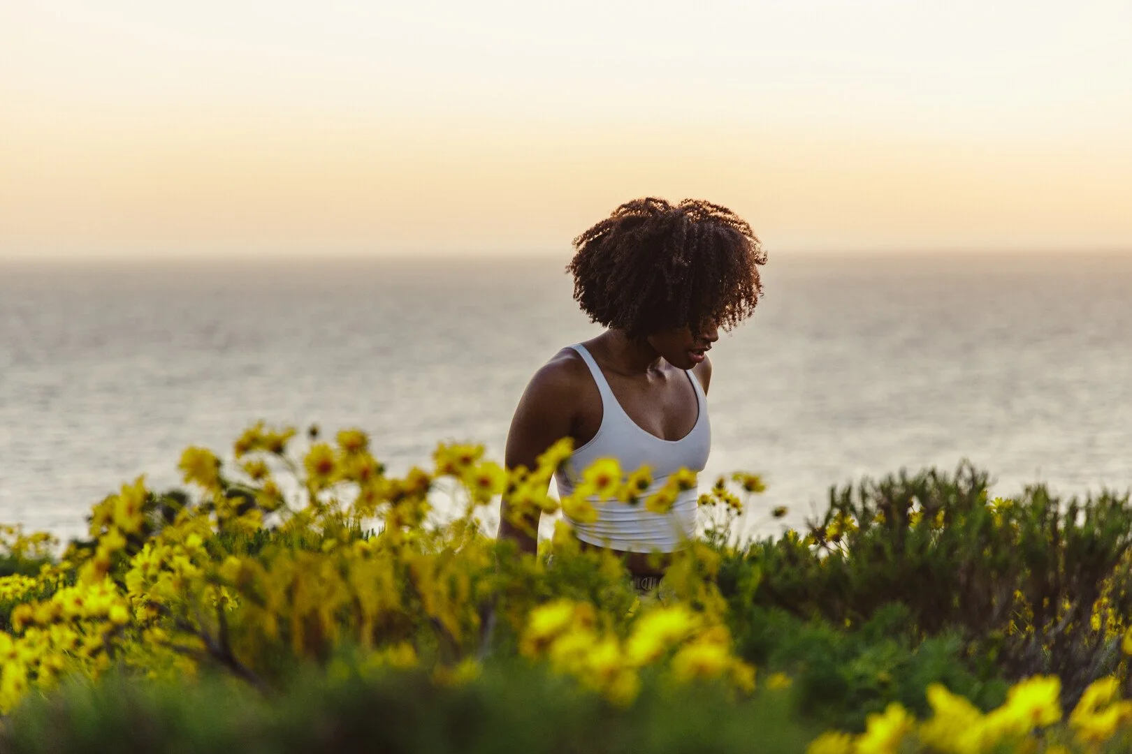 A woman with curly hair wearing a white tank top stands among yellow flowers on a hill, with the ocean and sunset in the background.