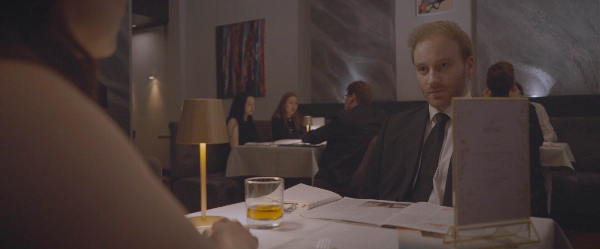 A man in a suit and tie sitting at a table in a dimly lit restaurant, with a glass of whiskey and a notebook in front of him, looking towards a woman across the table. Several other diners are visible in the background.