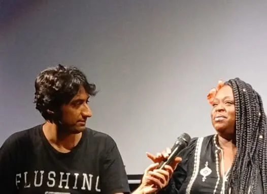 A young man in a black t-shirt holding a microphone on stage, listening to an older woman with braided hair speaking into the microphone at a panel or interview event.