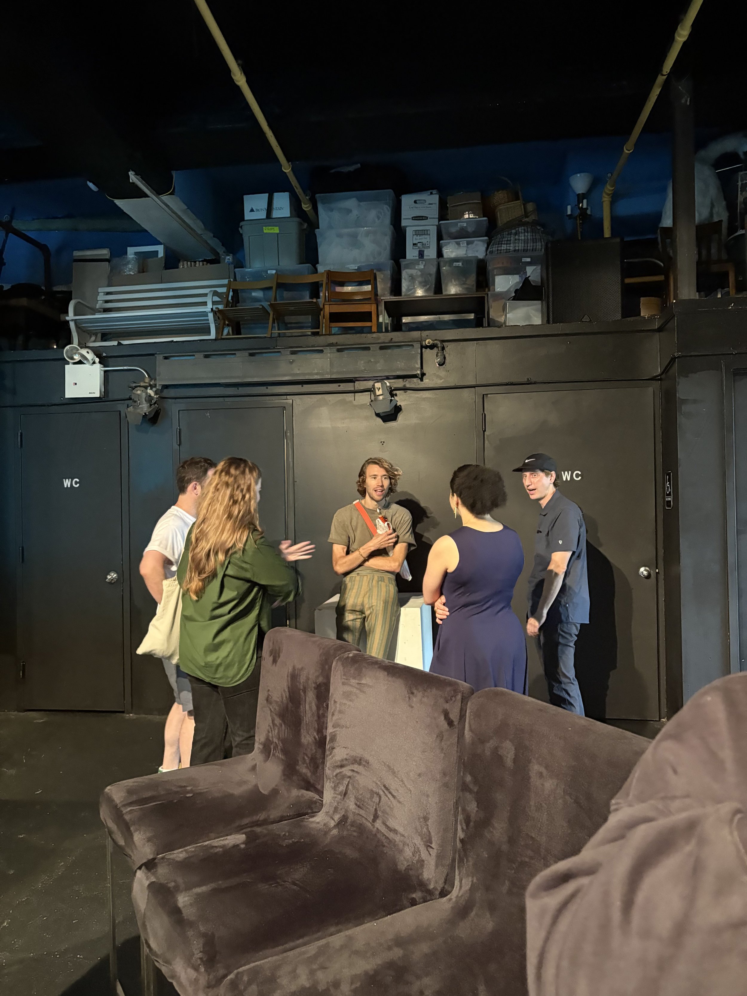 A group of five people having a conversation backstage of a theater or performance space, with a black wall marked 'WC' in the background, stacked chairs and storage items on a high shelf, and brown velvet furniture in the foreground.