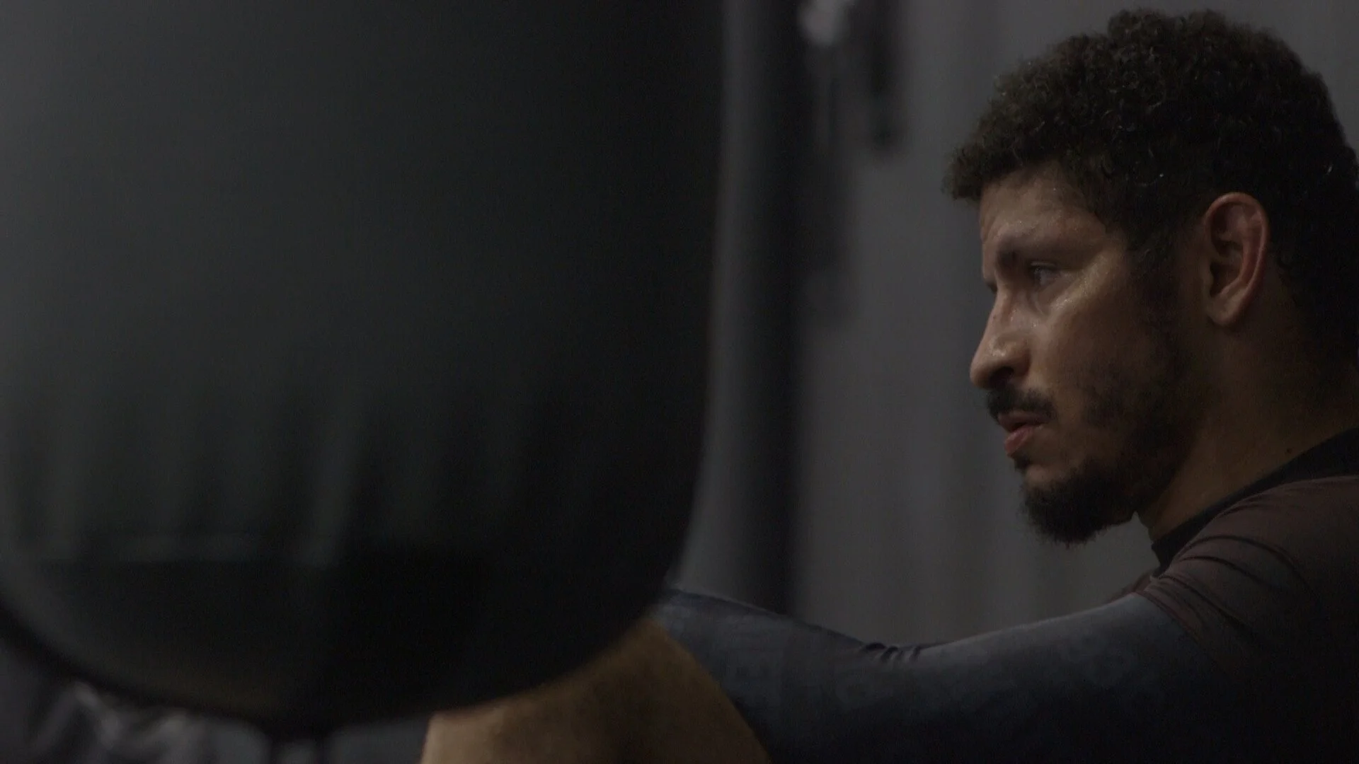 A man with curly hair and a beard, wearing a dark sports shirt, is sweating and appears to be resting or reflecting in a focused moment in a gym or workout environment.