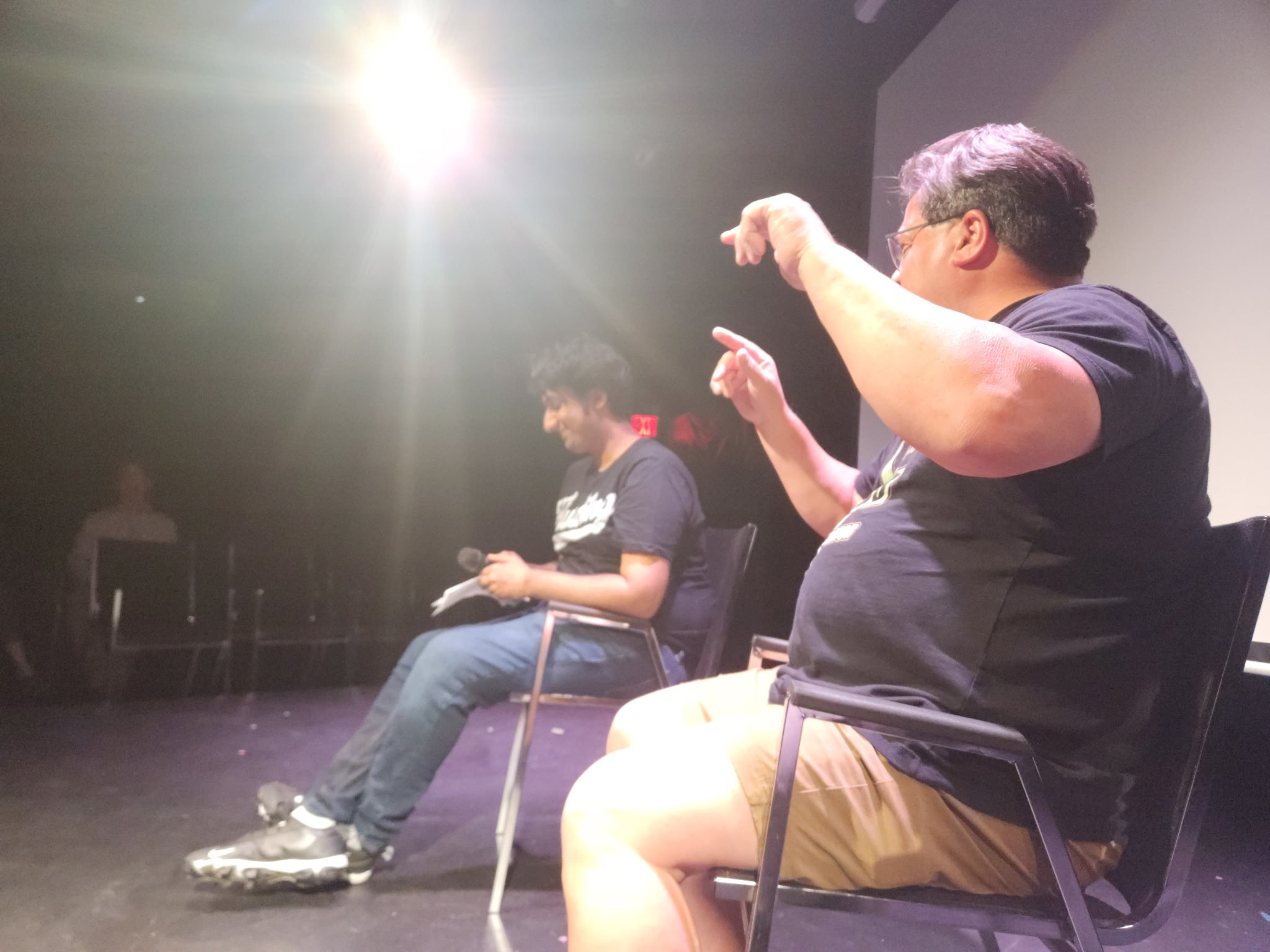 Two people seated on stage, one with a microphone and notes, and the other gesturing with their hands, under bright stage lighting.