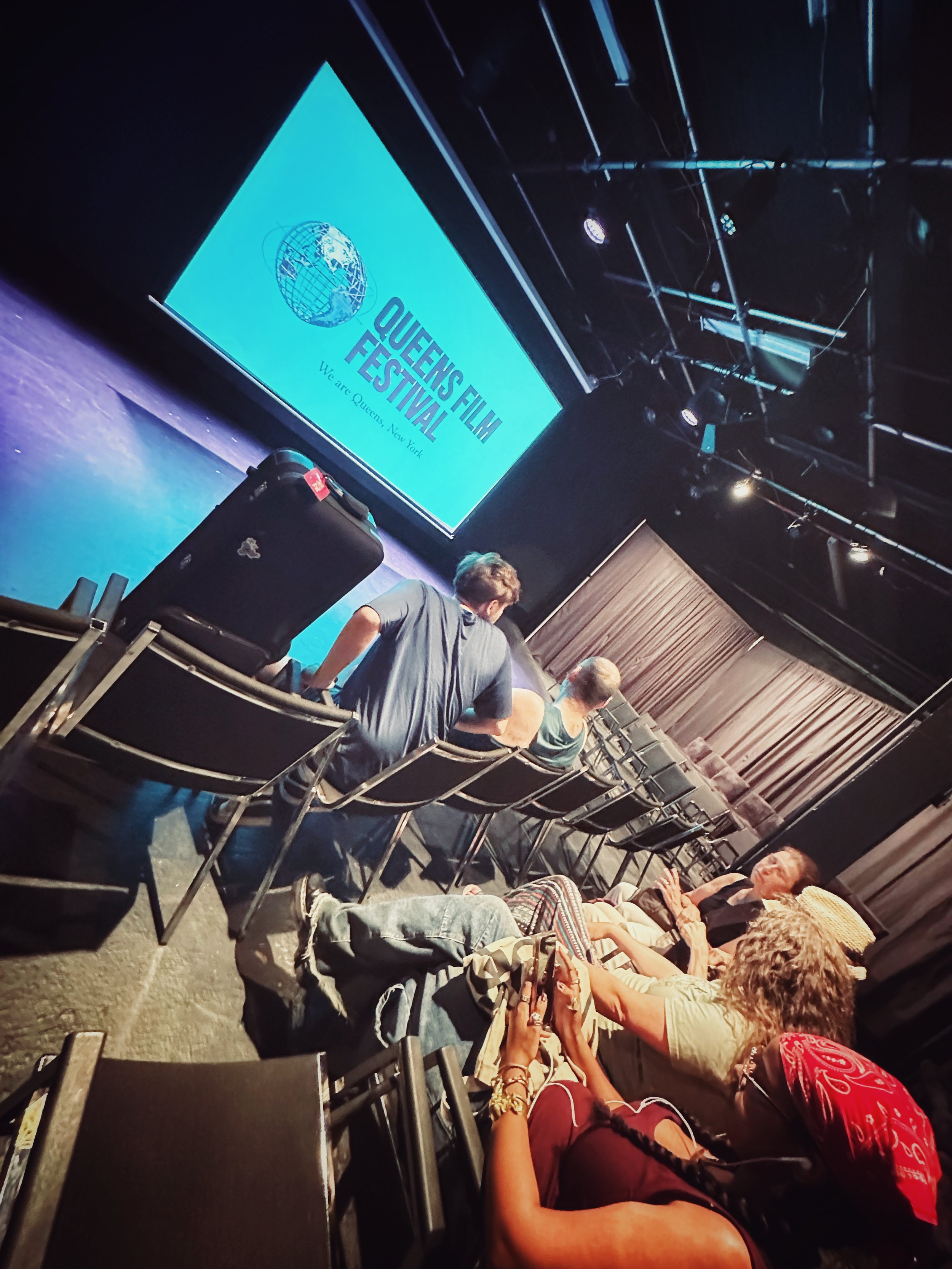 Audience seated in a theater at Queens Film Festival, watching a presentation on stage with a large screen displaying the Queens Film Festival logo and slogan, 'We are Queens, New York.'
