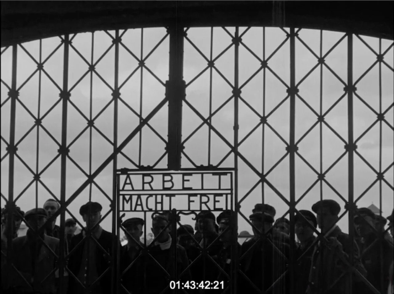 A black and white photograph of a group of people standing behind a metal gate with a sign that reads 'Arbeit Macht Frei' in German, meaning 'Work Sets You Free.'