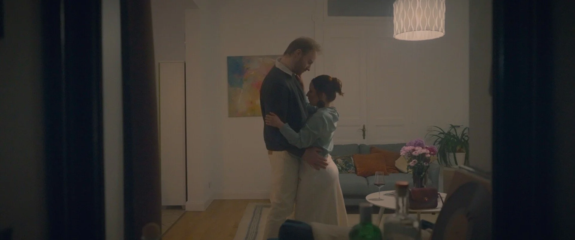 A man and woman share an intimate moment, embracing and smiling at each other in a warmly lit living room decorated with flowers and art.