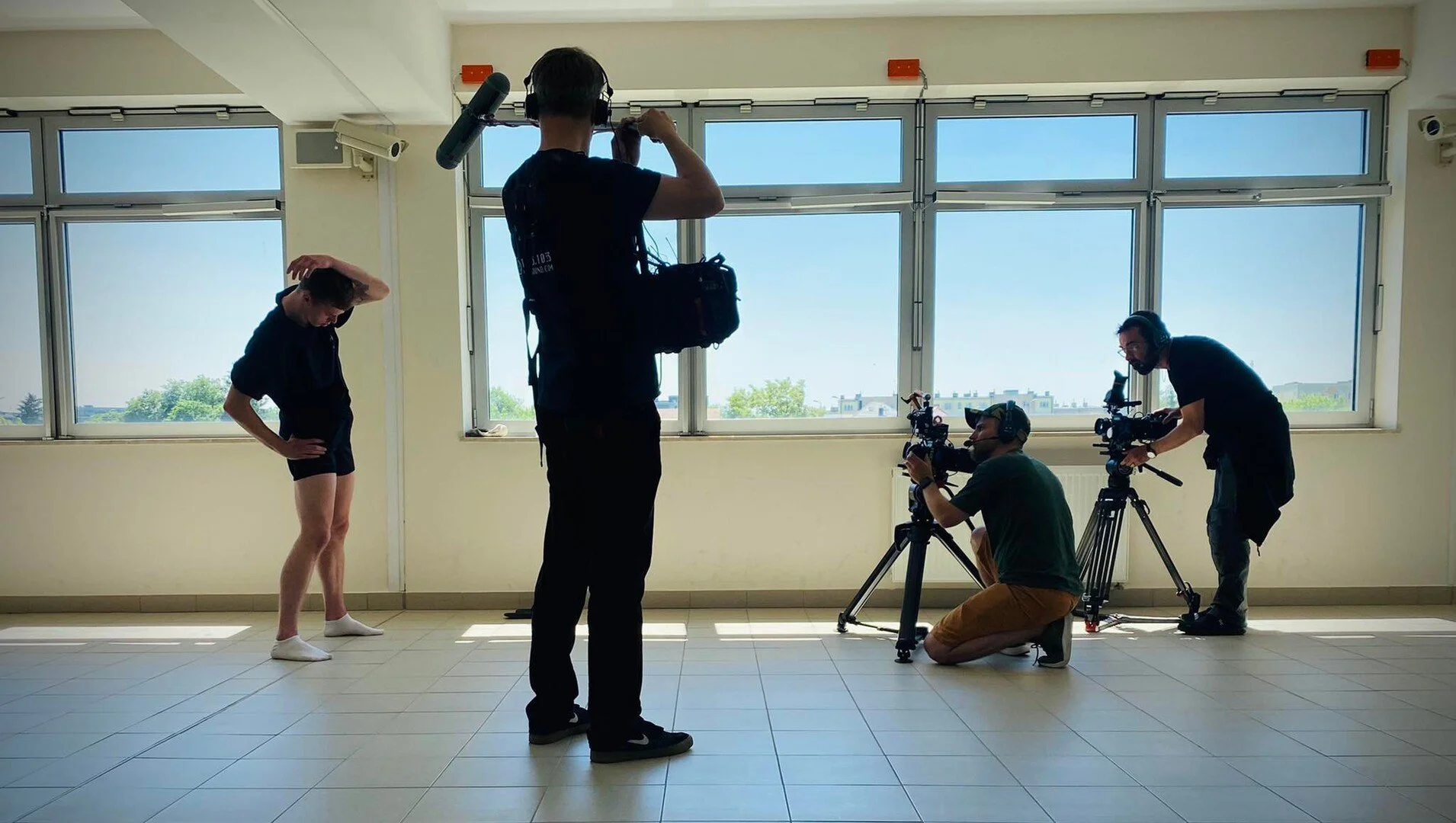 A filming crew is shooting a scene with a dancer in a bright room with large windows. The dancer is standing with one hand on her head, wearing black shorts and a black top. The crew members are operating cameras and equipment, and one person is adjusting his headphones.