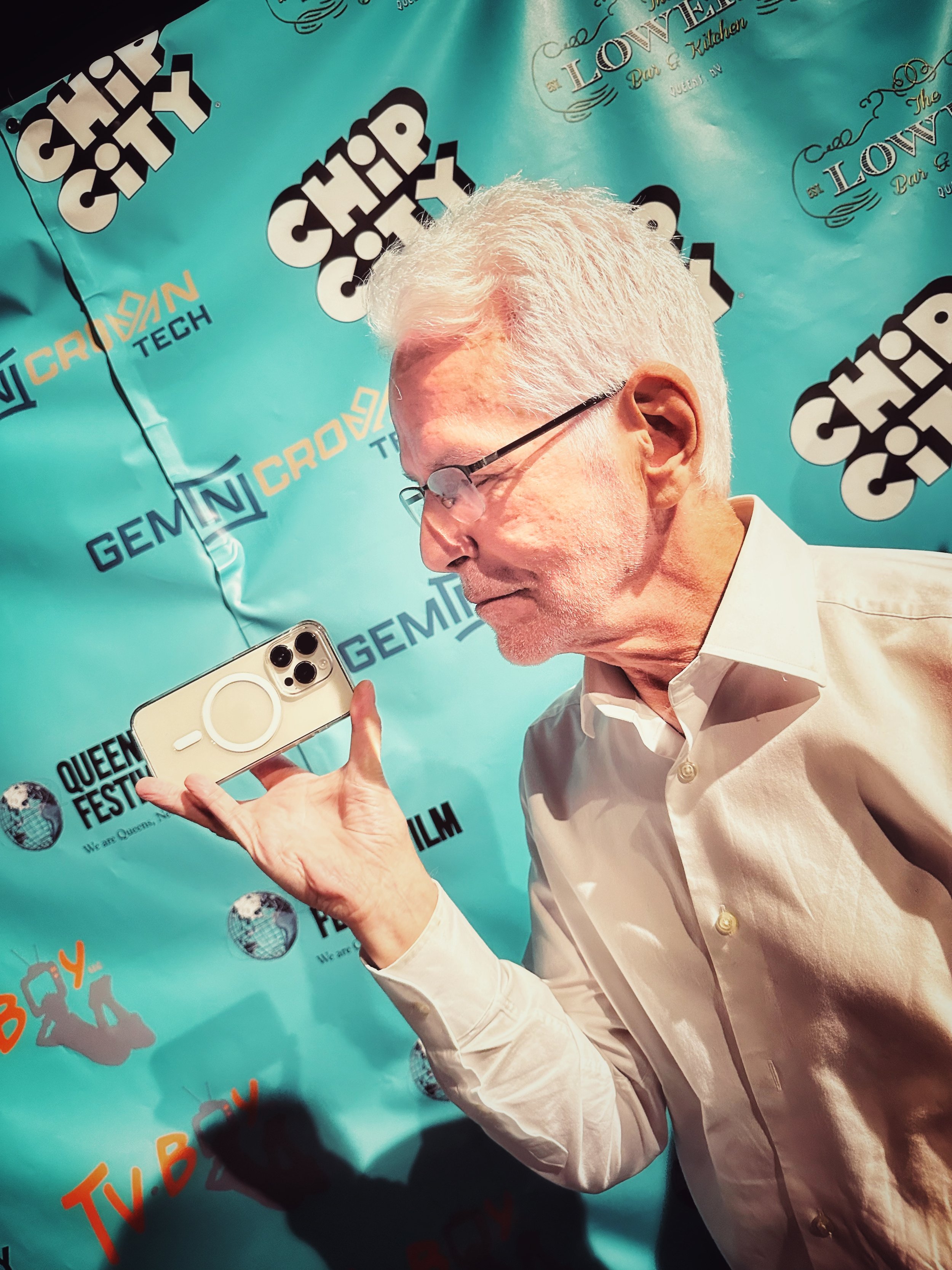 An elderly man with white hair and glasses holding a smartphone in front of a blue backdrop with event logos and texts, including 'GEMINI CROWN,' 'QUEEN FILM FESTIVAL,' and 'TV.BY'.