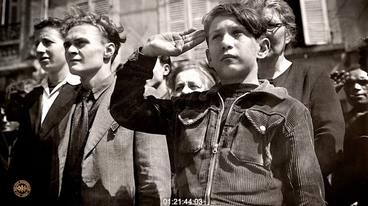 Black and white photo of a young boy saluting during a patriotic event, surrounded by adults and other children, in an urban area with older buildings in the background.