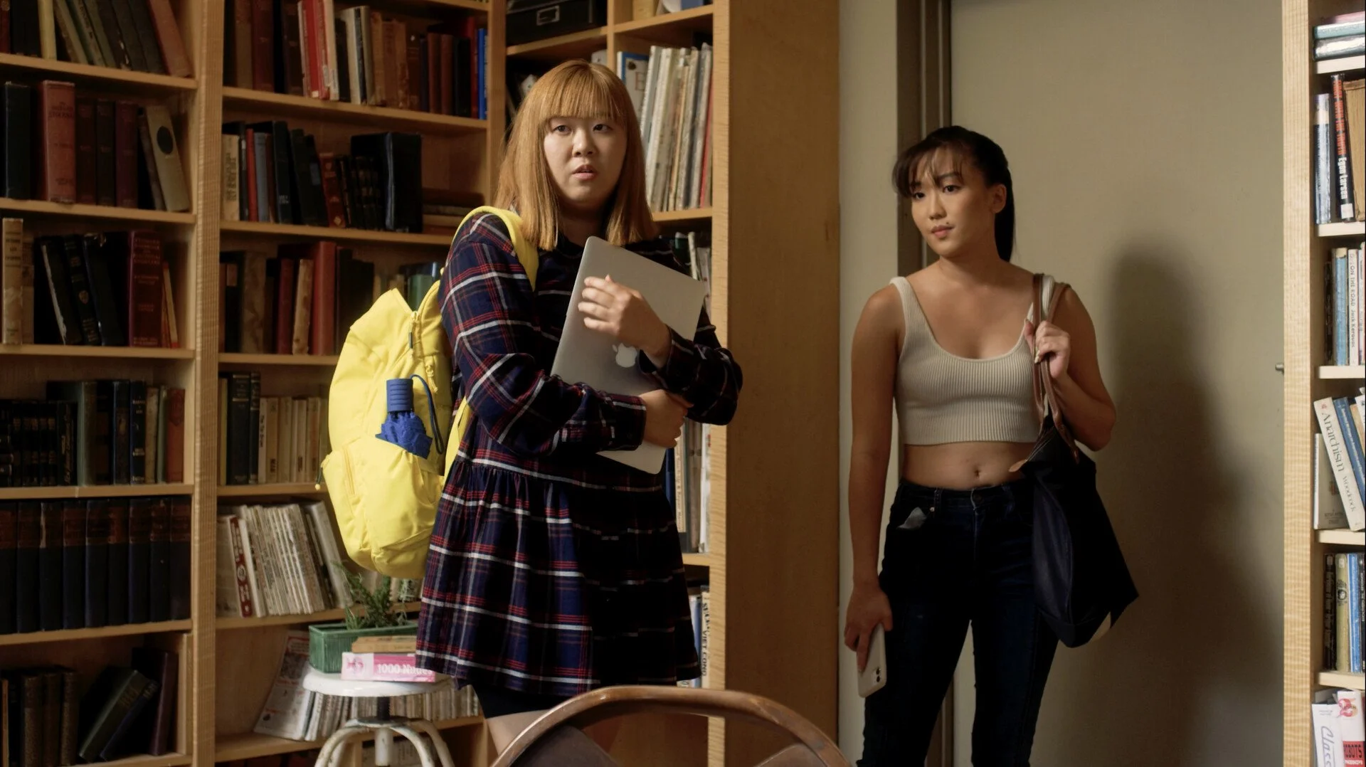 Two young women stand in a library. The woman on the left has red hair and is wearing a plaid dress with a yellow backpack, holding a laptop. The woman on the right has dark hair, a beige crop top, and jeans, holding a phone and a jacket.