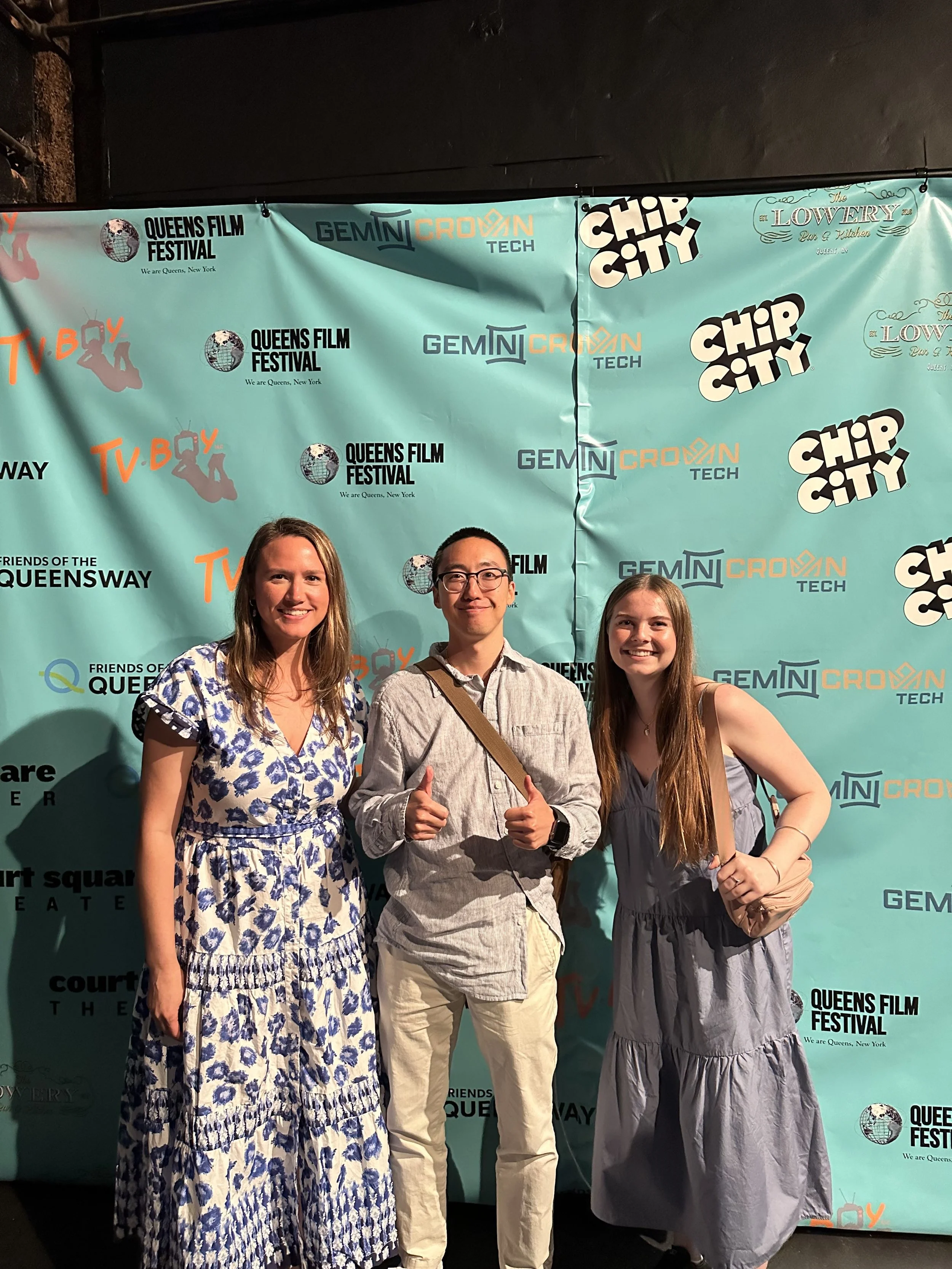 Three people standing in front of a teal backdrop with festival and event logos, smiling for the camera. The woman on the left wears a blue and white dress, the man in the middle wears glasses and a light-colored shirt with a brown strap, and the woman on the right wears a gray dress.