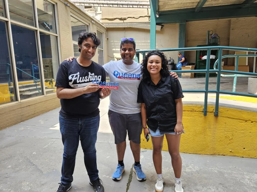 Three diverse smiling people standing together outdoors, with the two men on the left holding a sign that says 'Queens,' and the woman on the right holding a phone. Behind them is a woman sitting at a table under a covered area.
