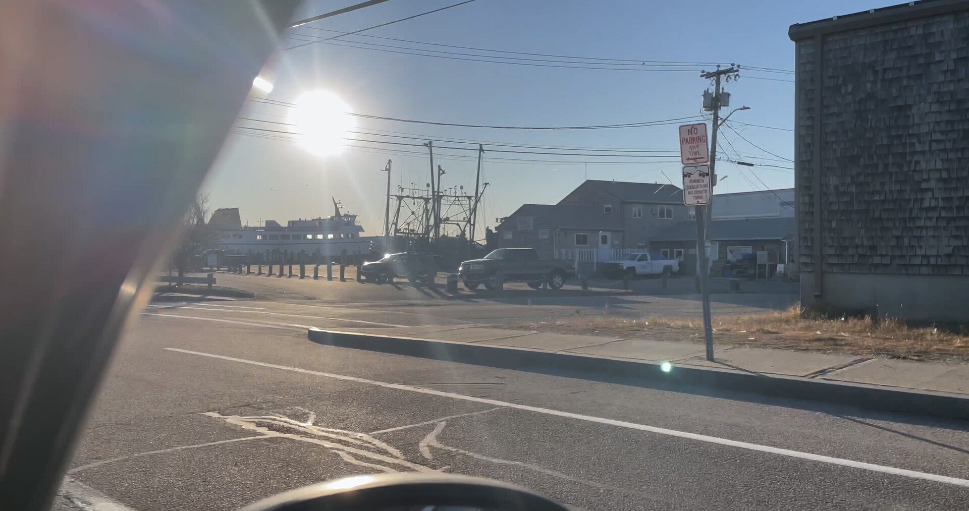 View of a harbor with boats, parked cars, houses, and utility poles with wires, taken from inside a vehicle with sunlight glare and a part of the vehicle's interior visible in the foreground.