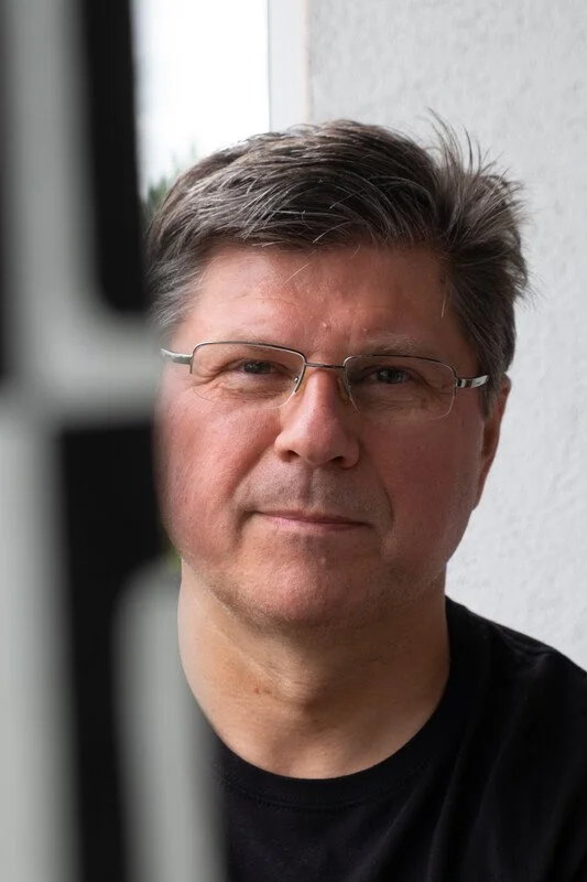 A middle-aged man with short brown hair, wearing glasses and a black t-shirt, looking at the camera with a neutral expression.