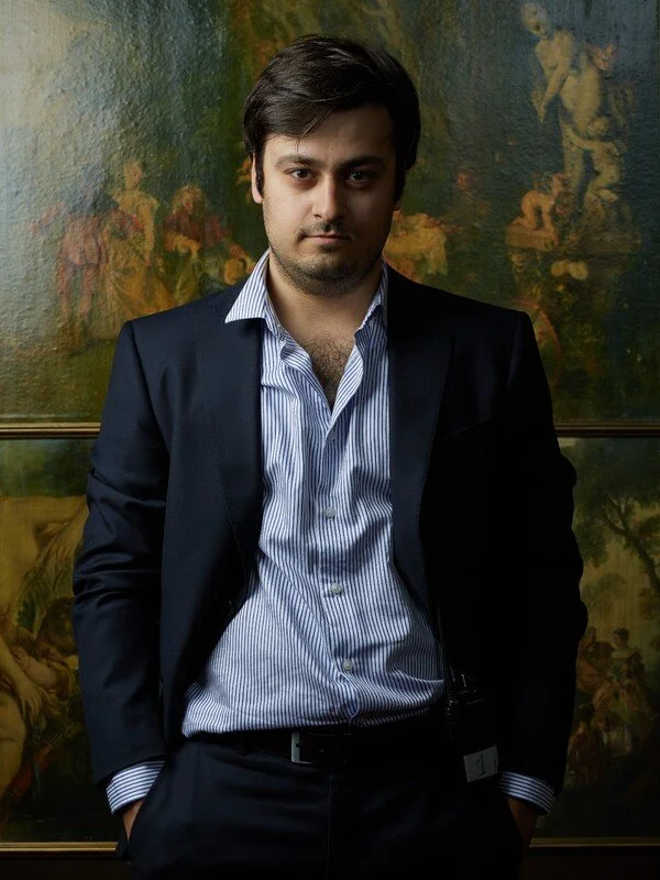 A young man in a dark suit with a light blue striped shirt, standing in front of a colorful painted background.