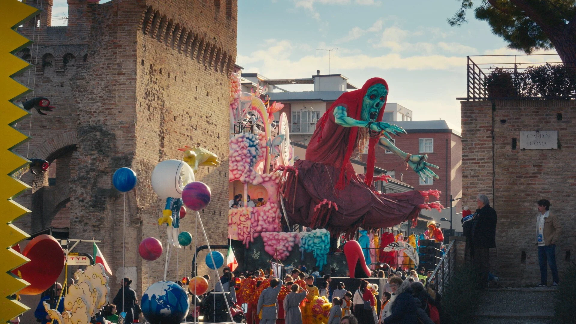A parade float featuring a large, moving puppet of a skeleton with a distorted face, dressed in red, surrounded by colorful decorations and balloons, with a crowd of people and historic brick buildings in the background.