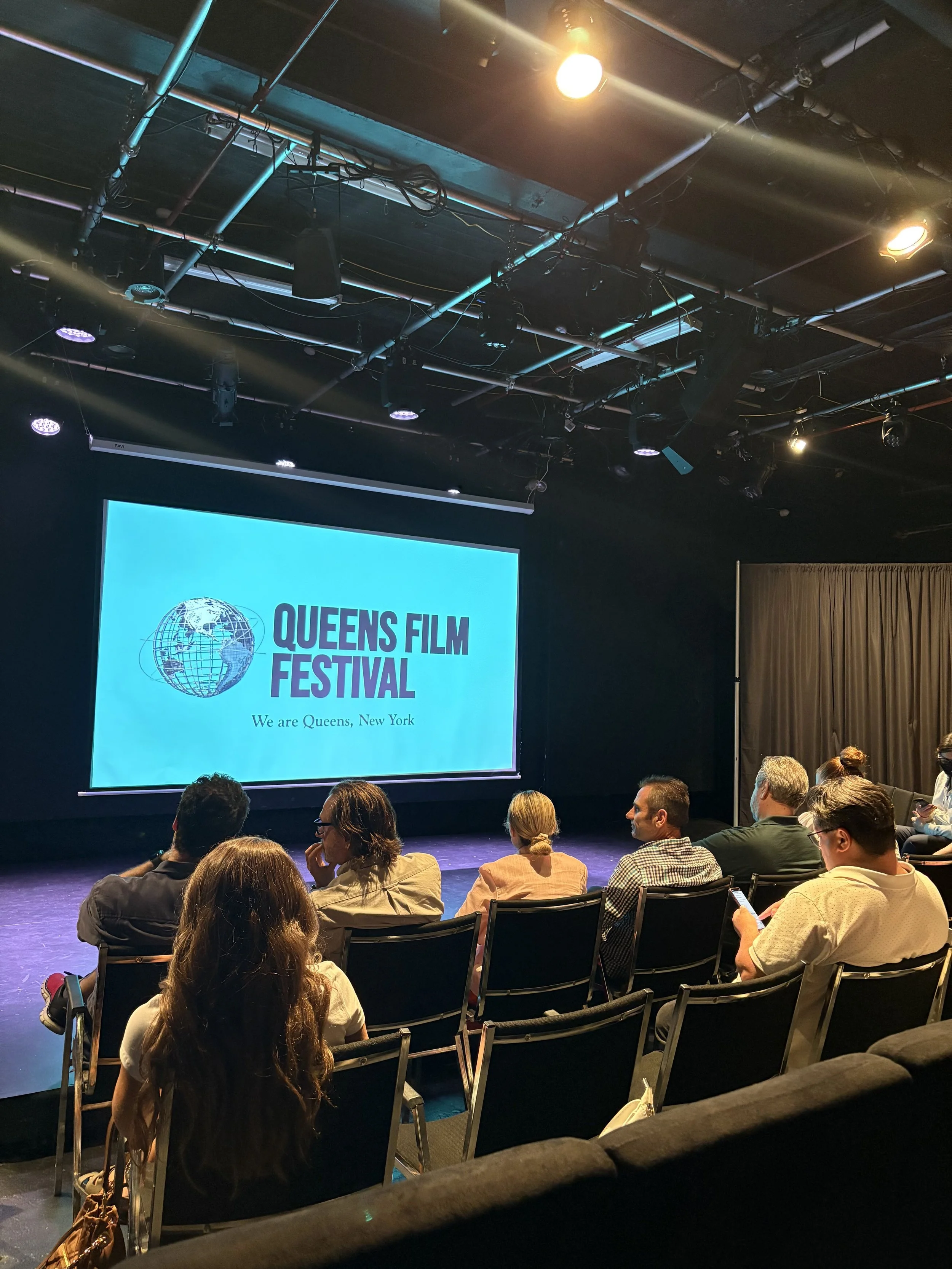 Audience sitting in a theater watching a presentation at Queens Film Festival, with a large screen displaying the festival's logo and tagline in Queens, New York.
