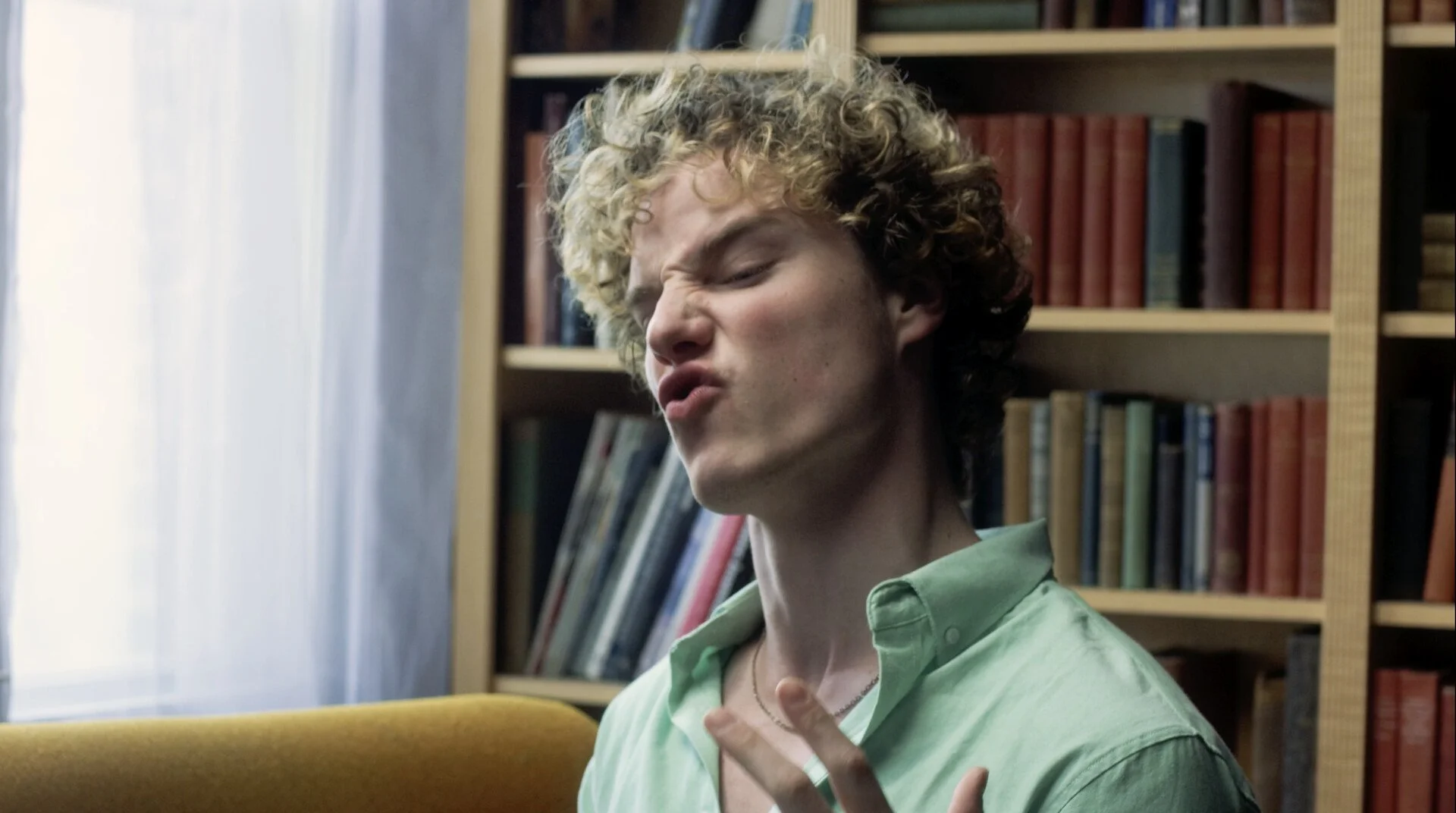 A young man with curly blonde hair making a facial expression of discomfort or pain, seated in a room with bookshelves in the background.