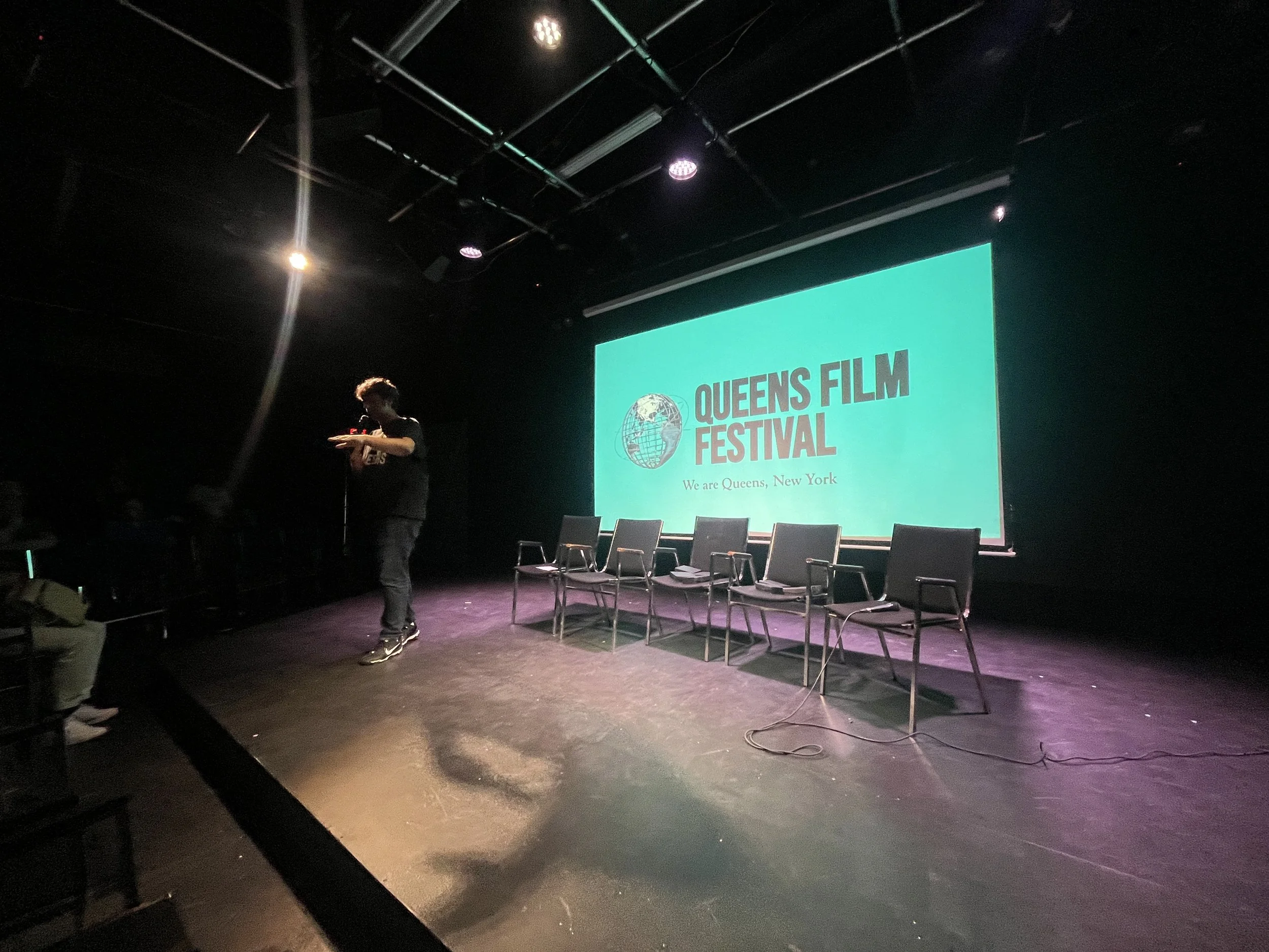 Stage with six empty chairs in front of an illuminated screen displaying the Queens Film Festival logo and the text 'We are Queens, New York.' A person stands to the left of the stage, looking at their phone in a dark auditorium.