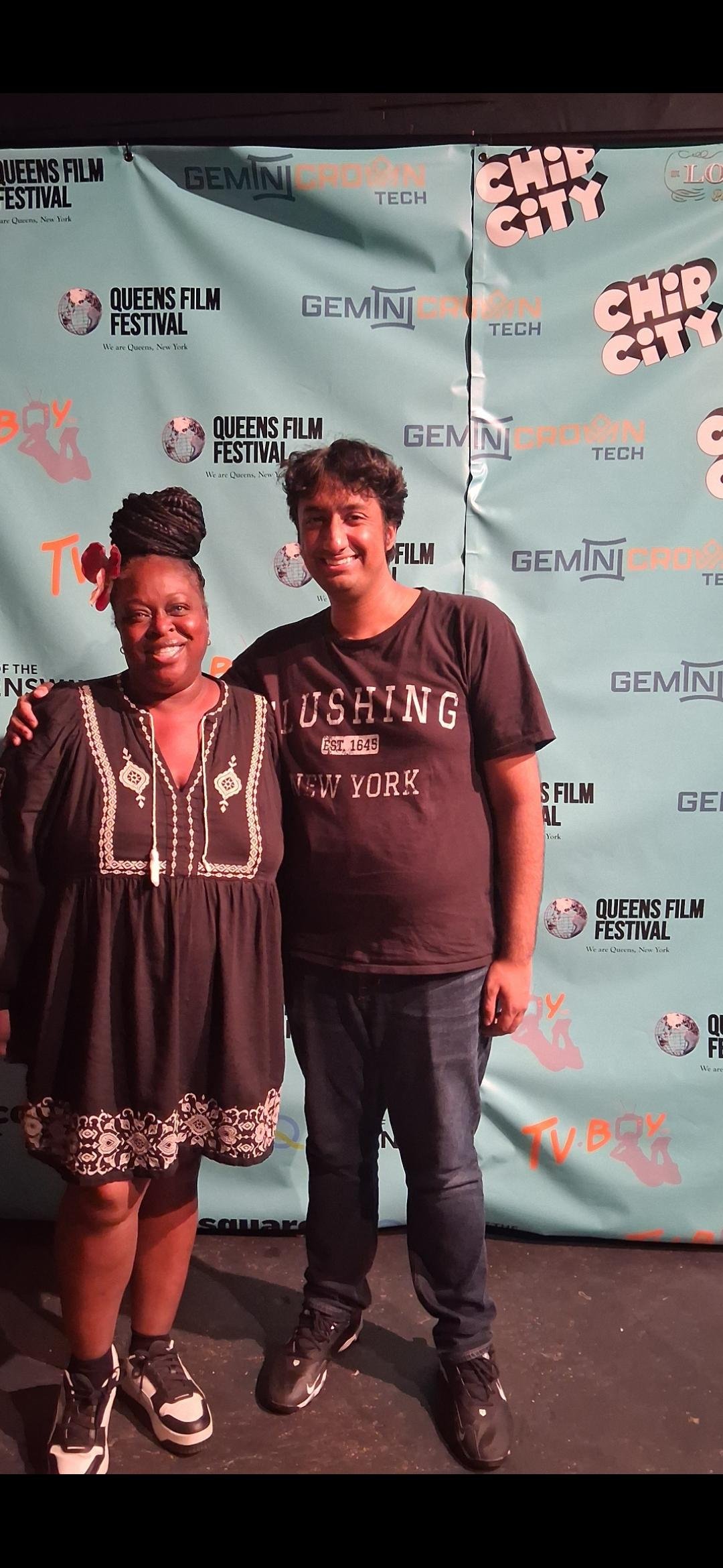 Two people posing for a photo in front of a backdrop with various logos and text, at the Queens Film Festival. The woman on the left is wearing a black dress with white embroidery and sneakers, and has her hair styled in braids piled on top of her head with a flower accessory. The man on the right is wearing a black T-shirt with white text, jeans, and sneakers.