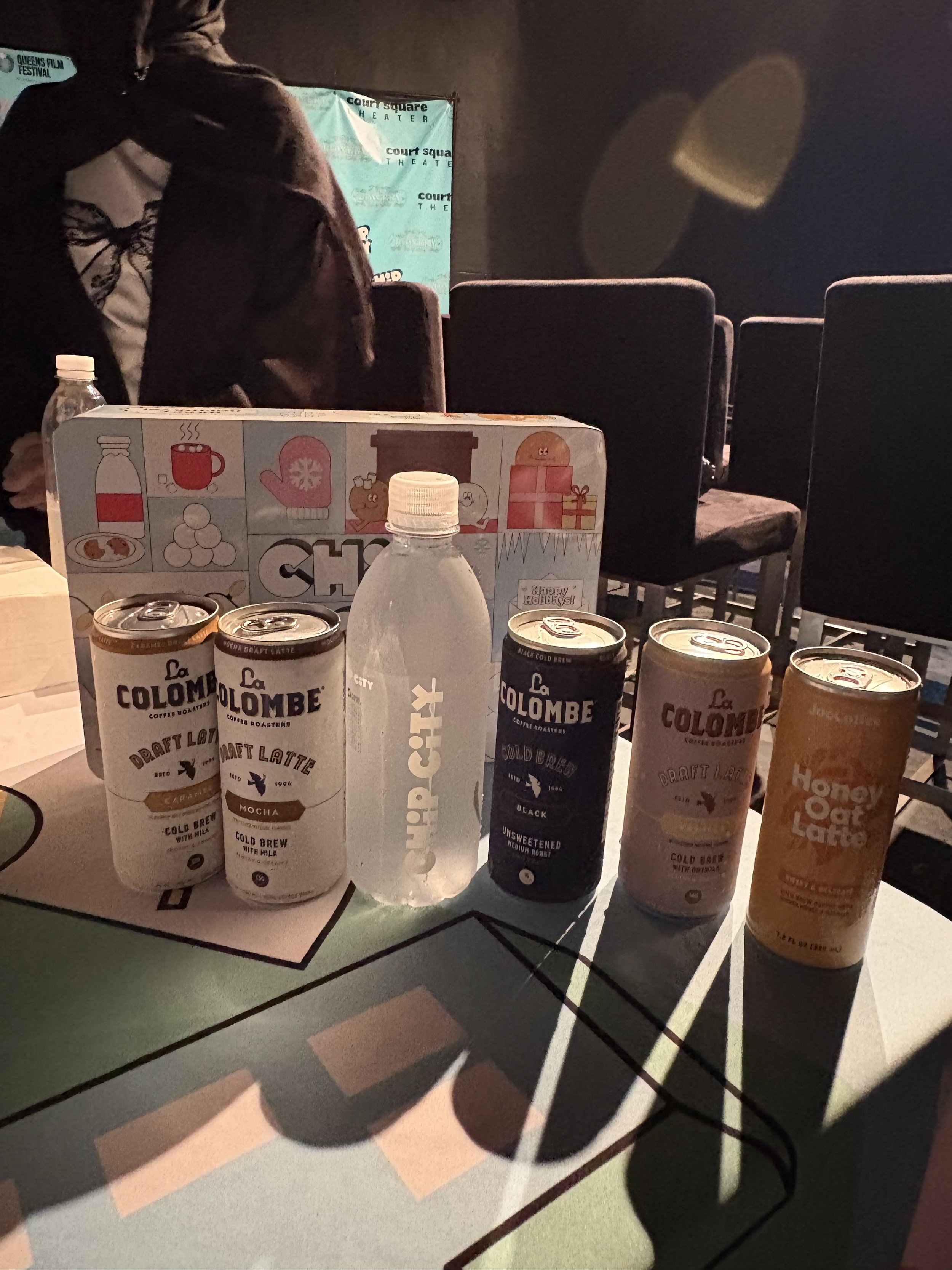 Table of canned La Colombe craft coffee drinks and a plastic bottle of Chick-fil-A water in a dark theater with empty chairs and a person standing in the background.