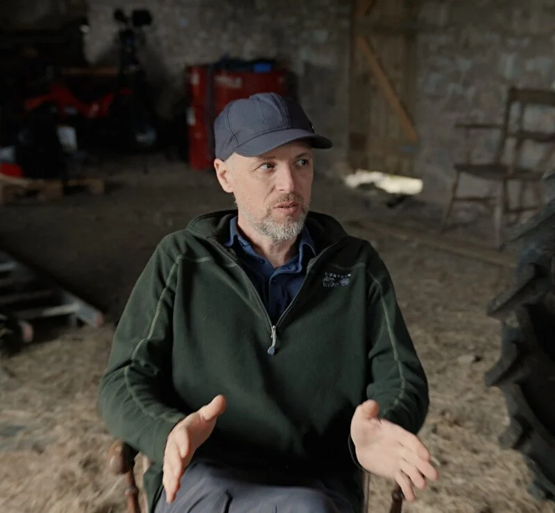 A man with a beard, wearing a dark cap and green jacket, sitting indoors in a rustic barn or shed.