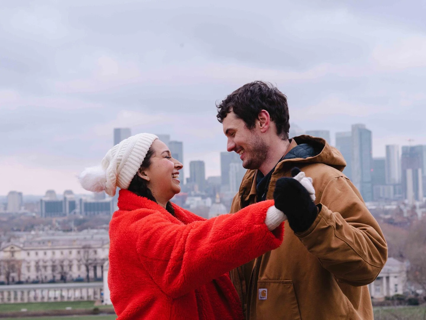 A smiling couple dancing outdoors with a city skyline in the background. The woman is wearing a red coat and a white knit hat, and the man is wearing a brown jacket. They are holding hands and looking at each other.