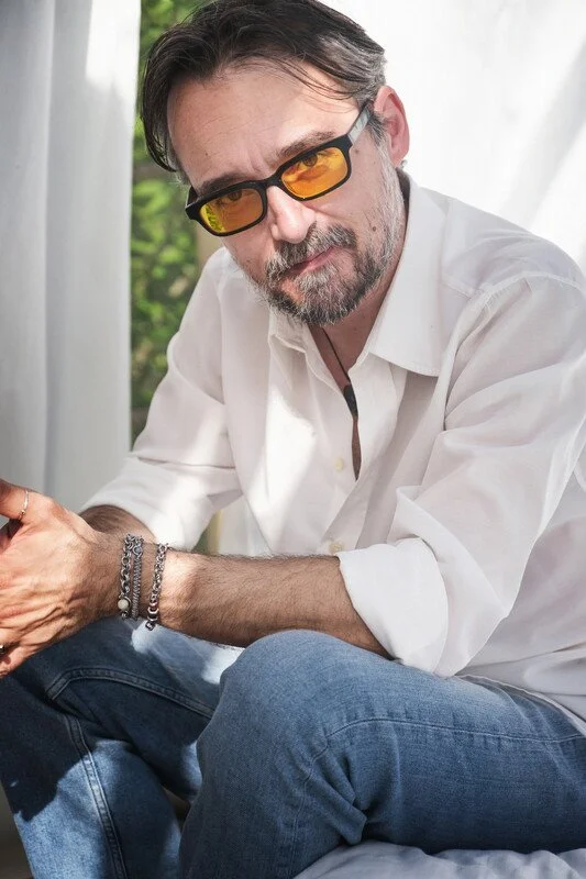 Man with dark hair, beard, and sunglasses with yellow lenses sitting by a window.