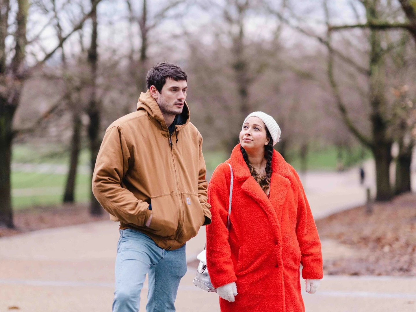 A man and woman walking outdoors in a park with leafless trees in the background. The man is wearing a brown jacket and jeans, while the woman is wearing a bright red coat, white beanie, and gloves.